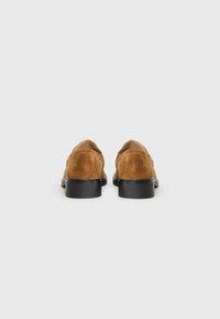 Des chaussures en daim marron avec des semelles noires vues de l'arrière, présentant un design simple et élégant sur un fond blanc.