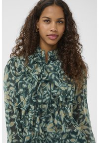 Fiatal nő göndör hajjal, teal színű fodros blúzt visel, amelyen levélminták láthatók, egyszerű fehér háttér előtt áll.