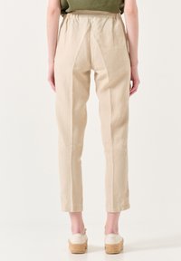 Beige Leinenhosen mit einer lockeren Passform, featuring einem elastischen Bund, sichtbaren Nähten und schmal zulaufenden Beinen, kombiniert mit hellen Schuhen.