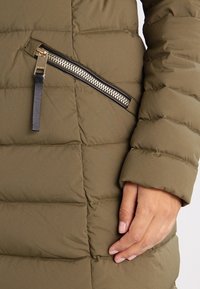 Olivově zelená prošívaná bunda s šikmou kapsou na zip a rukou opřenou o manžetu rukávu.