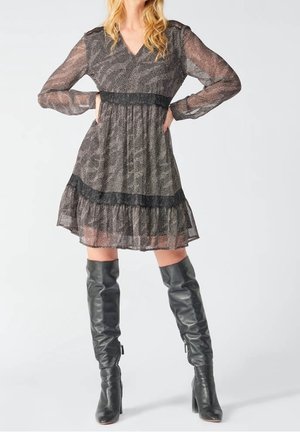 Kobieta w przezroczystej, wzorzystej sukience z długim rękawem, ozdobionej czarną koronką, oraz w czarnych skórzanych, sięgających do kolan butach na obcasie, stojąca z rękami na biodrach.