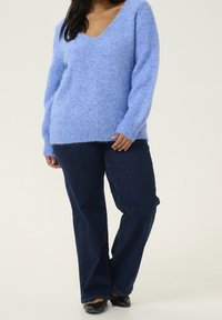 Pull en maille pelucheux bleu clair avec un col en V profond, associé à un jean droit bleu foncé et des chaussures plates noires. Texture douce, coupe décontractée.