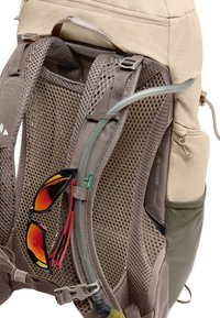 Sac à dos beige avec panneau arrière en maille. Ses caractéristiques incluent des bretelles ajustables, un tuyau d'hydratation et des lunettes de soleil rouges et noires attachées.