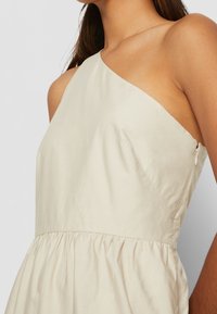 Vestido asimétrico de color crema con un corsé ajustado y una falda recogida y fluida. Hecho de tela liviana con cierre de cremallera lateral.