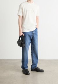 Muž v bílém tričku Calvin Klein Jeans, modrých džínách, černých botách, držící malou černou kabelku přes rameno, stojící na betonové podlaze.