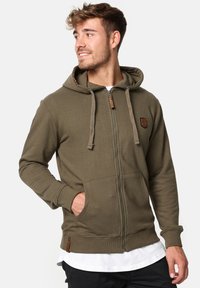Veste à capuche zippée olive en tissu doux, dotée d'une capuche avec cordon de serrage, de poches avant et d'un accent en patch texturé.