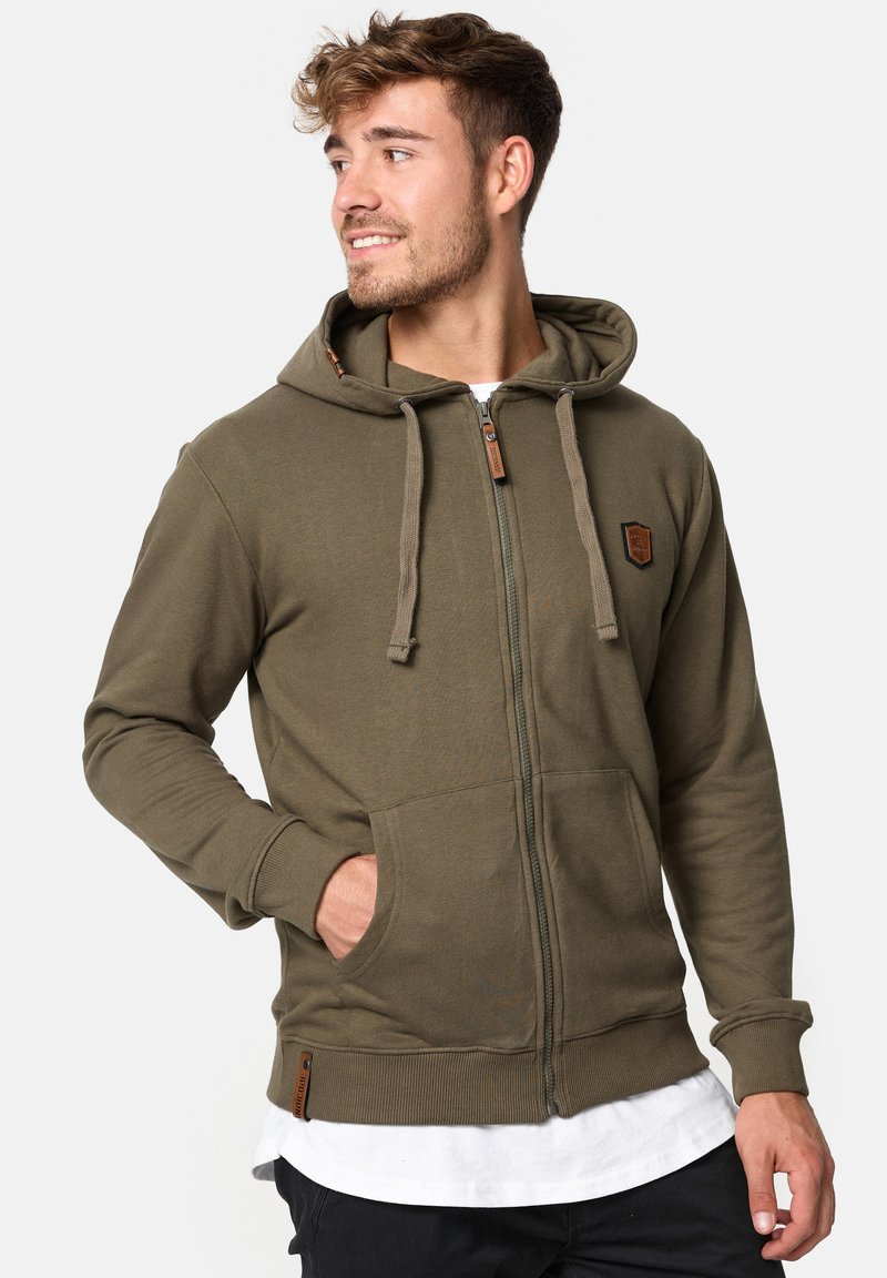 Veste à capuche zippée olive en tissu doux, dotée d'une capuche avec cordon de serrage, de poches avant et d'un accent en patch texturé.