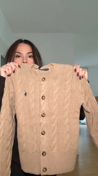 Beige stickad kofta med flätmönster, rund halsringning och fem stora bruna knappar. Liten logotyp på vänster sida.