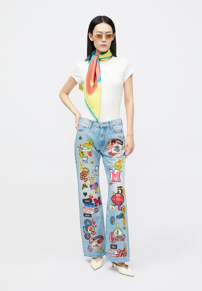 Femeie îmbrăcată cu un tricou alb, blugi largi de un blue deschis cu pete colorate, o eșarfă galbenă cu model, pantofi cu toc bej și ochelari de soare amber, în picioare.