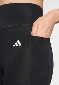 Sorte atletiske leggings lavet af strækbart stof, med en sidelomme og hvidt Adidas-logo. Glat tekstur med flade sømme.