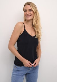 Juodas be rankovių marškinėlis su V formos iškirpte, pagamintas iš lygaus audinio. Modelis dėvi šviesiai mėlynus džinsus, laikydamas viršutinės dalies apačią.