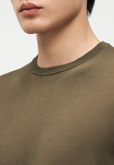 Κοντινό πλάνο στο κάτω μέρος του προσώπου και το λαιμό ενός ατόμου που φοράει πουλόβερ με λαιμόκοψη σε ελιά χρώμα, πάνω σε απλό φόντο.