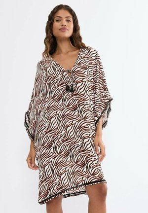 Woman wearing a loose, knee-length dress with a brown and black zebra print and V-neck tied with tassels, standing against a plain background.