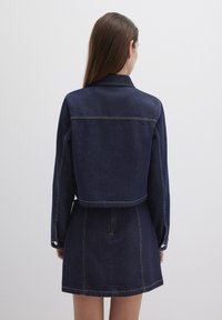 Denim jacket with a cropped design, dark blue color, and yellow stitching details. Features long sleeves and snap button closures.