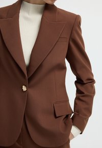 Blazer marrone su misura realizzato in tessuto liscio, presenta un rever a punta, chiusura con un bottone e una vestibilità slim. Il rivestimento interno mostra una texture a coste.