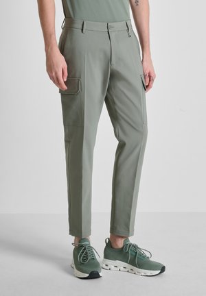 Uomo che indossa pantaloni cargo verde oliva slim fit con tasche con patta e scarpe da ginnastica verdi abbinate, in piedi contro uno sfondo grigio chiaro uniforme.