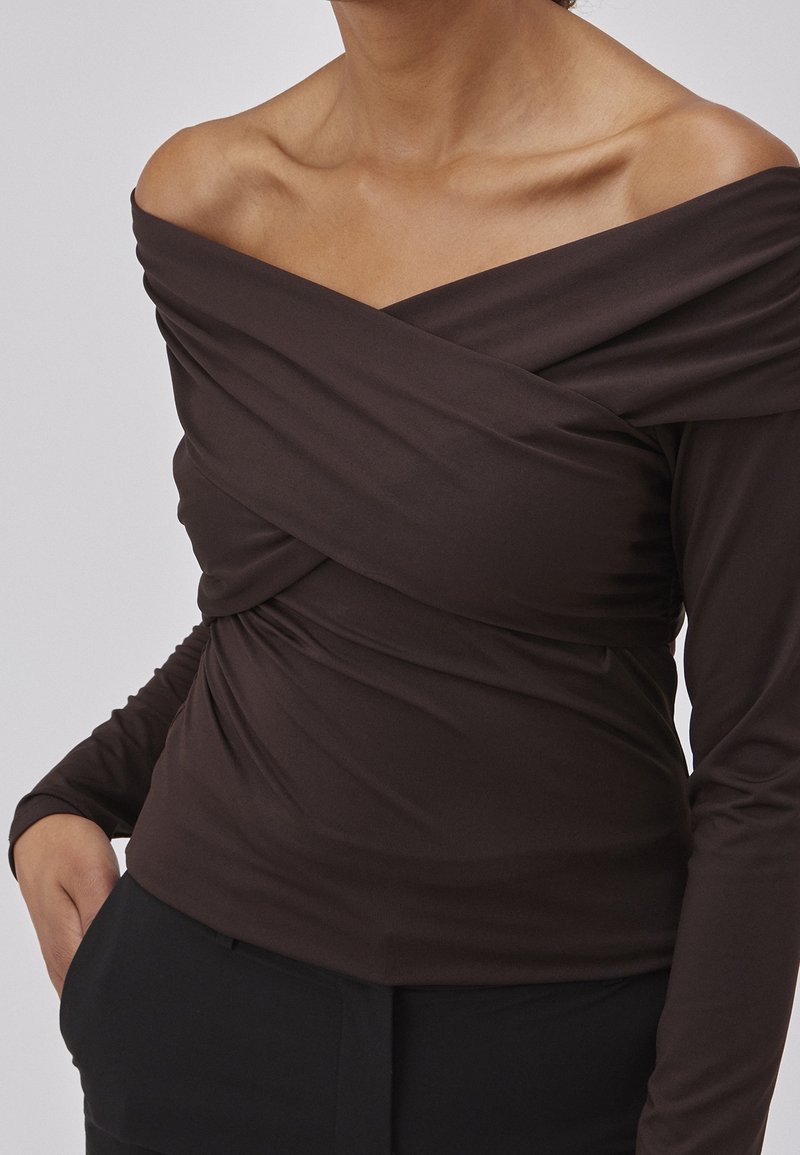 Personne portant un haut moulant à manches longues, épaules dénudées, de couleur marron foncé avec un détail de tissu croisé au niveau de la poitrine et un pantalon noir.