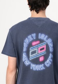 Blå bomull T-skjorte med et rundt grafisk motiv på baksiden med teksten "CONEY ISLAND NEW YORK CITY" og fargerike designelementer.