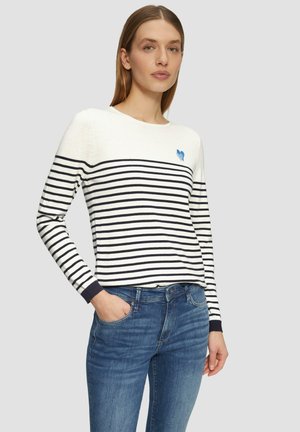Maglione a righe con maniche lunghe in bianco e navy, dotato di un piccolo emblema a forma di cuore blu sul petto. Abbinato a jeans blu.