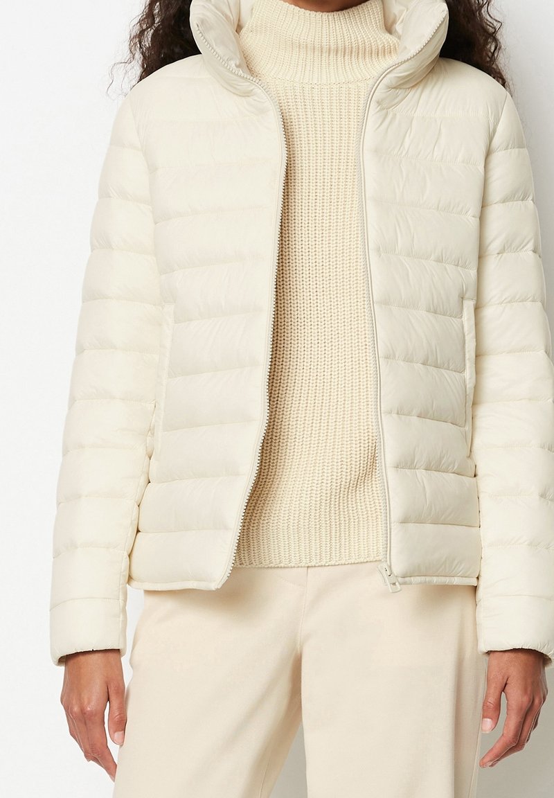 Femme portant une doudoune crème sur un pull à col roulé crème texturé et un pantalon crème assorti, debout devant un fond uni.