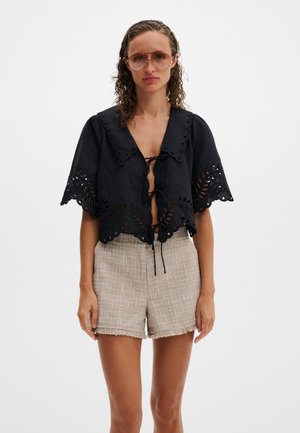 Sieviete ar cirtainiem matiem valkā lielizmēra tonētās saulesbrilles, melnu īss piedurkņu topu ar caurumiem un saitēm, kā arī bēšas, teksturētas augsta vidukļa šortus.