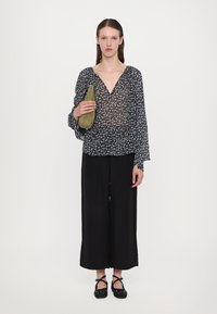 Blouse noire légère à motif floral avec un décolleté en V, associée à un pantalon large noir et un sac à main vert. Chaussures noires à boutons.