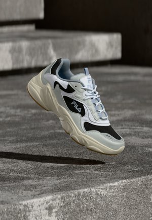 Zapatilla FILA blanca y negra con suela gruesa, flotando sobre escalones de hormigón con su sombra visible debajo.