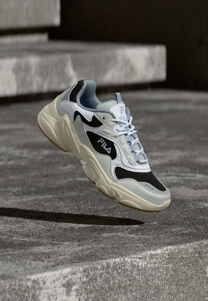 Λευκό και μαύρο αθλητικό παπούτσι FILA με χοντρή σόλα, αιωρούμενο πάνω από τσιμεντένια σκαλοπάτια με τη σκιά του να διακρίνεται από κάτω.