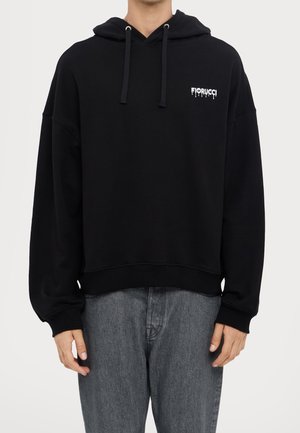 Mand iført sort Fiorucci hoodie med snore og lille hvidt logo på brystet, kombineret med grå jeans, mod en ensfarvet baggrund.
