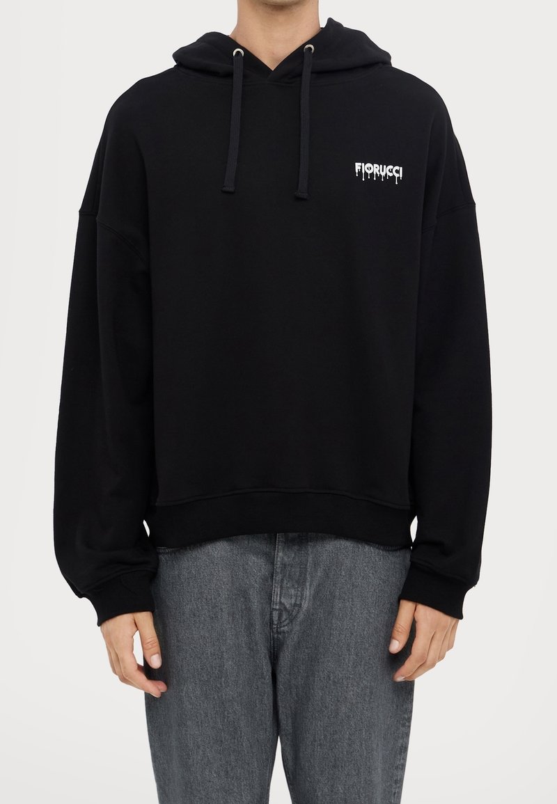 Homme portant un sweat à capuche noir Fiorucci avec cordons et petit logo blanc sur la poitrine, associé à un jean gris, sur fond uni.