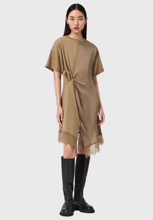 Frau mit langen dunklen Haaren, die ein taupefarbenes asymmetrisches Kleid mit Spitzenabschluss und schwarze kniehohe Stiefel trägt, steht vor einem schlichten Hintergrund.