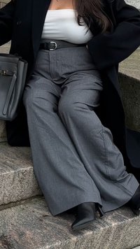 Femme assise sur des marches en pierre portant un pantalon gris à jambes larges, une ceinture noire, un manteau noir, un haut blanc, des bottes noires et tenant un sac à main noir.