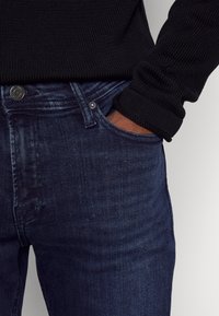Jeans de ganga de cor azul-escuro com uma textura subtil. As características incluem um design clássico de cinco bolsos e fecho de botão metálico na cintura.