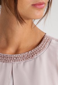 Blouse rose clair présentant un col rond orné de perles légères texturées et d'un corps en tissu lisse. Fini avec un subtil éclat.