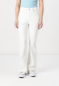 Person wearing high-waisted white flare jeans and white platform sneakers, standing against a plain white background.