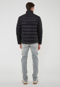 Giovane uomo che indossa un giubbotto nero imbottito, jeans azzurri chiari e sneakers bianche, in piedi, di spalle, contro uno sfondo bianco semplice.