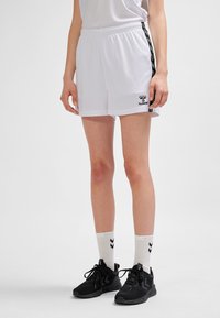 Weiße Sportshorts mit schwarzen Seitenstreifen, elastischem Bund und einem kleinen Logo auf der linken Seite. Kombiniert mit schwarzen Turnschuhen und weißen Socken.