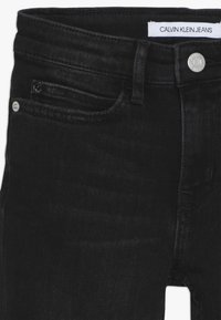 Jeans de mezclilla negra con corte recto, que cuentan con un cierre de botón plateado, bolsillos frontales y una etiqueta con el sello "Calvin Klein Jeans".