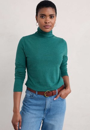 Zelený rolák z měkkého materiálu, s dlouhými rukávy a volným střihem, kombinovaný s modrými džíny s vysokým pasem a hnědým opaskem.