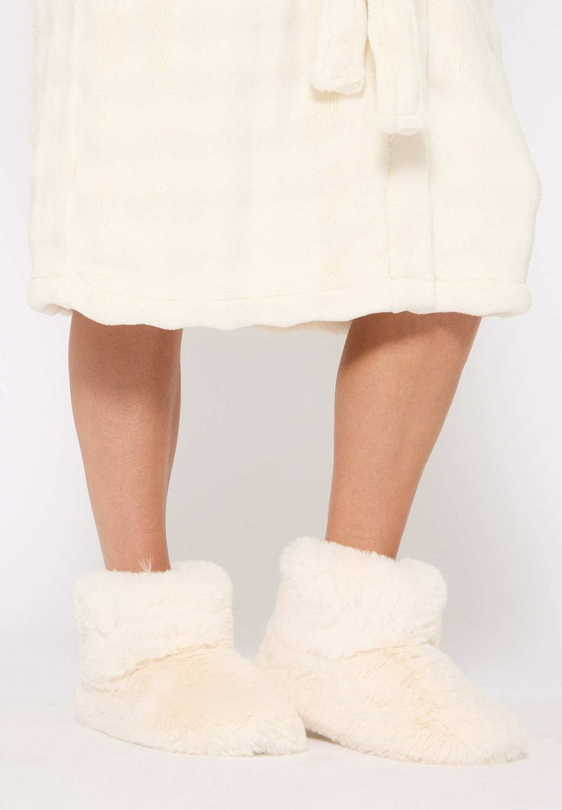 Zapatillas de felpa blancas con una suave textura de piel, que presentan un diseño de puño doblado y punta redondeada, complementando un albornoz esponjoso a juego.