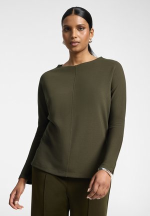 Femme aux cheveux foncés attachés, portant un haut à manches longues olive et un pantalon, des boucles d’oreilles créoles argentées et un bracelet argenté au poignet gauche.