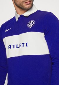 Blå och vit rugbystil skjorta med krage, med horisontell rand, "ATLETI" text och logotyp på vänster bröst. Blandning av bomullstextur.