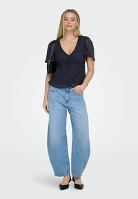 Femme portant un haut en dentelle bleu marine à manches courtes, un jean large bleu clair, des chaussures slingback noires et un sac à épaule noir, debout avec une main dans la poche.