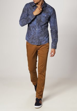 Hombre caminando con una camisa azul de botones con estampado, pantalones marrones y zapatillas negras con suelas blancas.