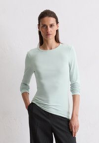 Helles mintgrünes Langarmoberteil mit glatter Textur und Rundhalsausschnitt, kombiniert mit dunkel gestreiften Hosen. Einfaches, körpernahes Design.