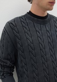 Grauer gestrickter Pullover mit einem gerippten Rundhalsausschnitt, der ein erhabenes Zopfmuster und eng anliegende Ärmel aufweist. Strukturiertes Gewebe mit subtiler Sheen.
