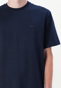 T-shirt en coton bleu marine avec un col rond et des manches courtes. Présente un logo noir brodé sur la poitrine gauche. Texture lisse.