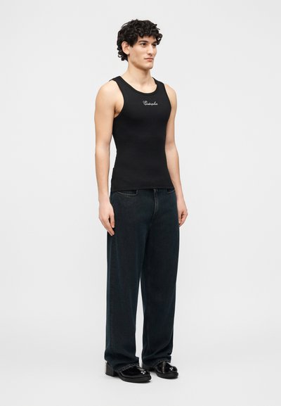 Jeune homme aux cheveux bouclés portant un débardeur noir avec le texte "Endorphin", un jean sombre ample et des chaussures noires brillantes, debout devant un fond blanc.