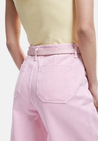 Pantalones rosa de tiro alto con una tela de mezclilla texturizada, que presentan dos bolsillos traseros y un cinturón a juego, combinados con una blusa de color amarillo claro.
