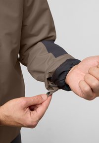 Waterdichte jas met een bruine hoofdkleur, donkergrijze accenten, elastische manchetten en een drukknoopsluiting op de mouw.